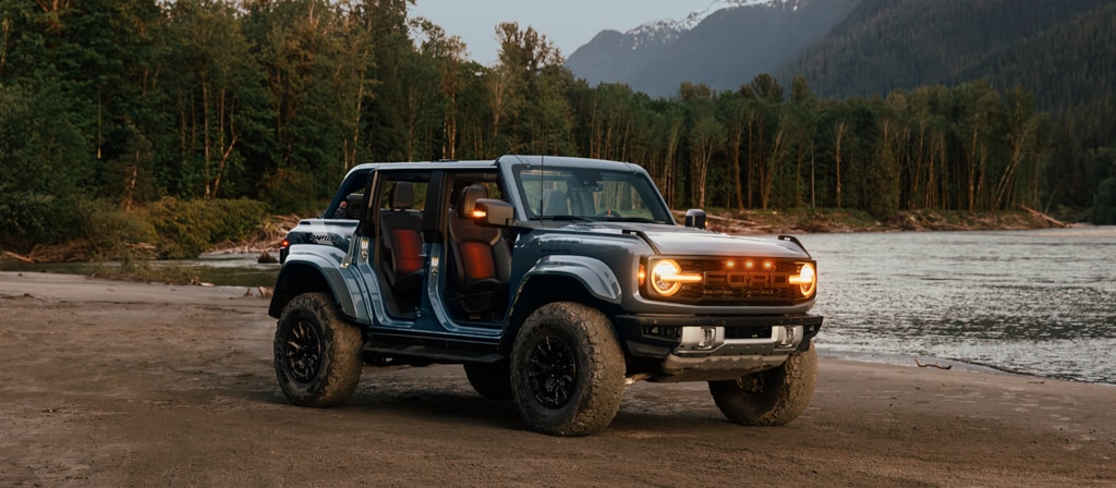 A 2025 Ford Bronco® SUV with the doors off and lights on, parked near a lake in the forest