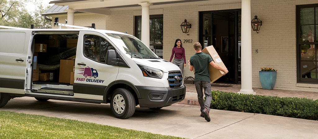 A white 2025 Ford E-Transit™ van parked as its driver walks to a house for delivery