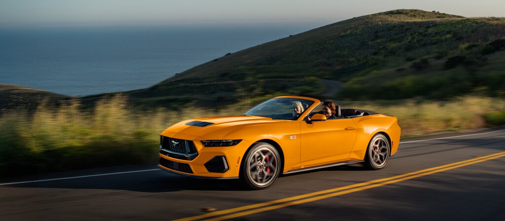 Two people in a 2026 Ford Mustang® GT convertible in Orange Fury Metallic Tri-coat being driven on a coastal highway