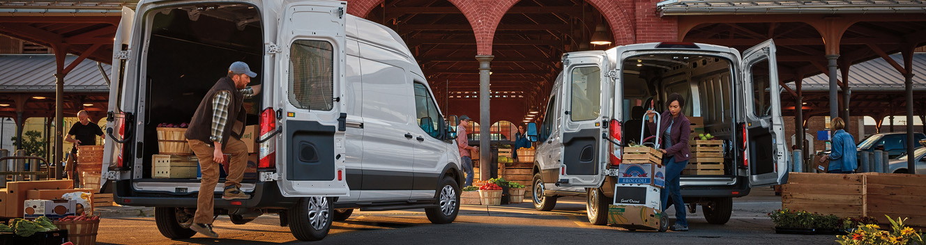 Two white 2027 Ford Transit® Vans with rear open and people unloading produce at a market