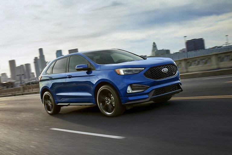 A Blue Ford Edge drives along a highway with a city skyline in the background