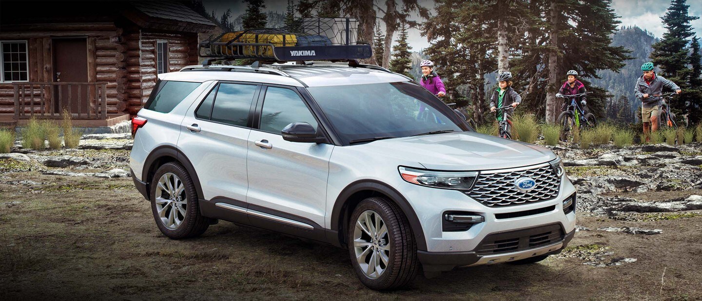 A family of four is biking towards a white 2023 Ford Explorer parked outside a cottage in a forest setting