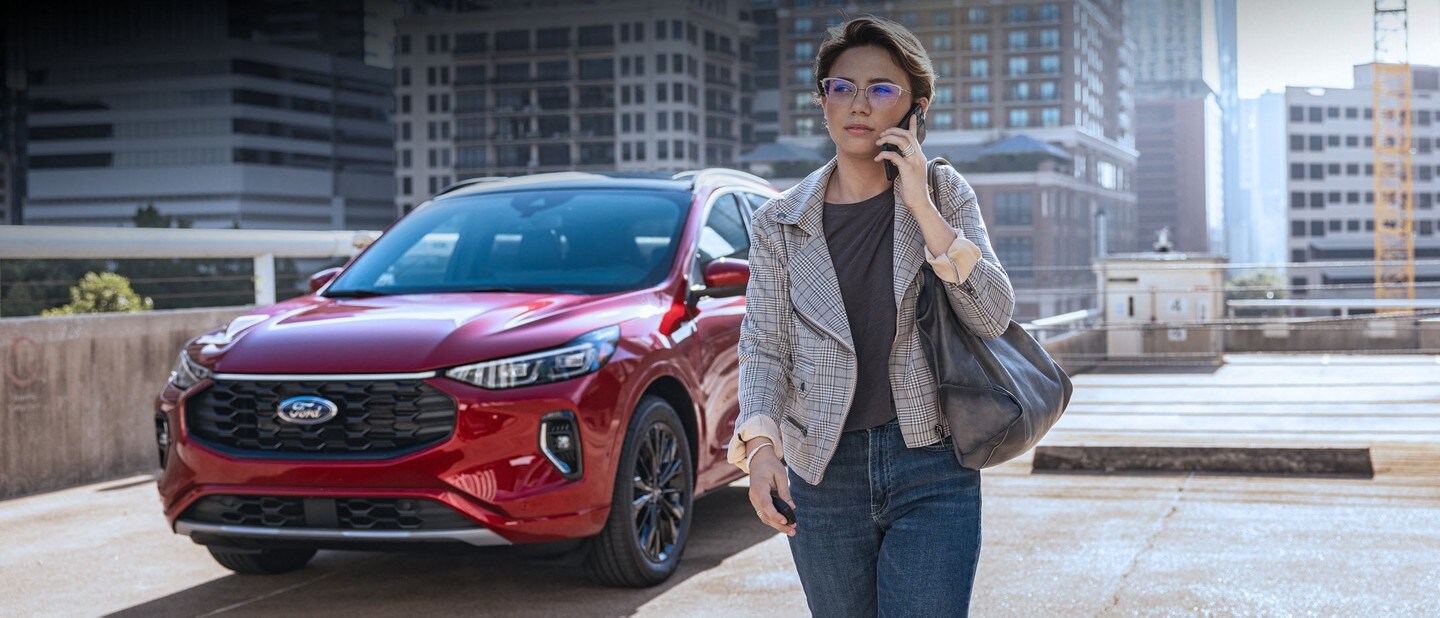 A woman talks on the phone as she walks away from a red 2023 Ford Escape parked on the roof of a parking garage.
