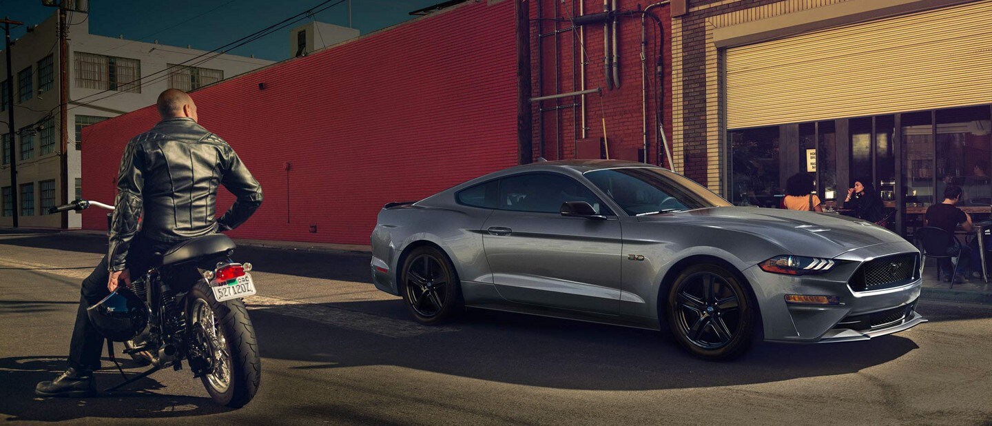 A man is sitting on his motorcycle while looking at a 2023 Ford Mustang in Carbonized Grey outside of a café