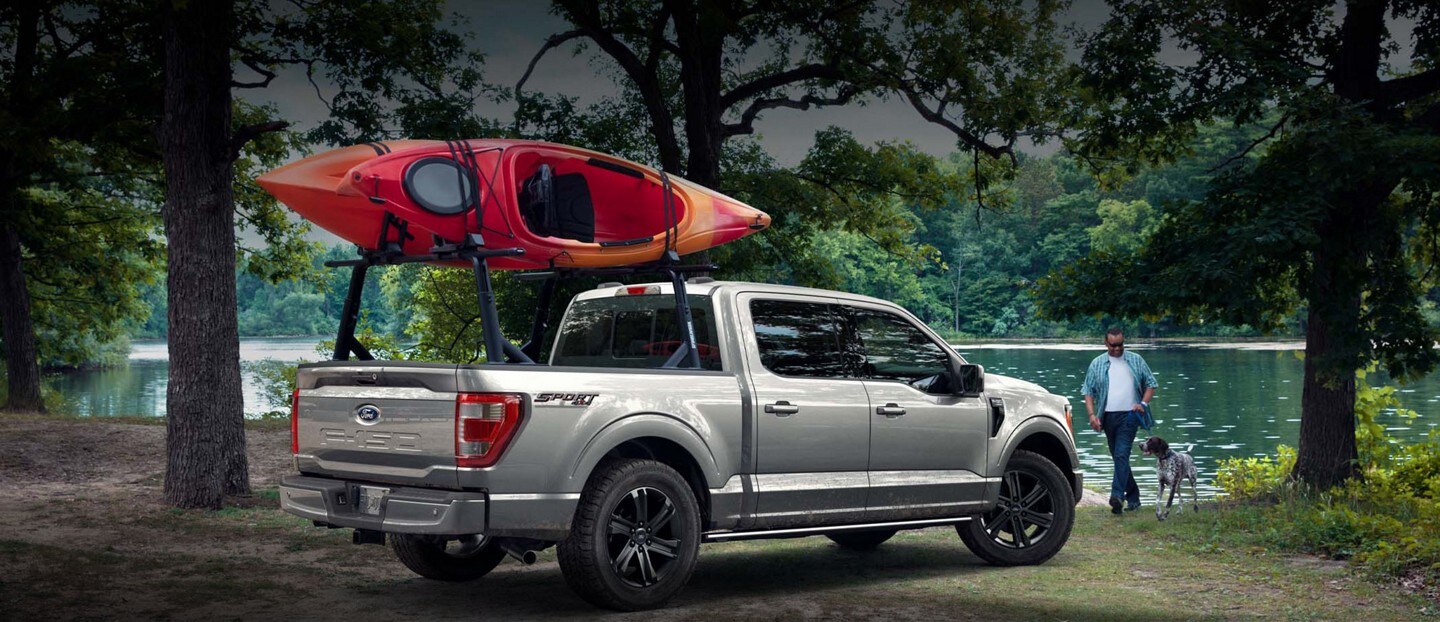 A man and his dog walking towards a silver 2023 Ford F-150 holding a red kayak by a lake.
