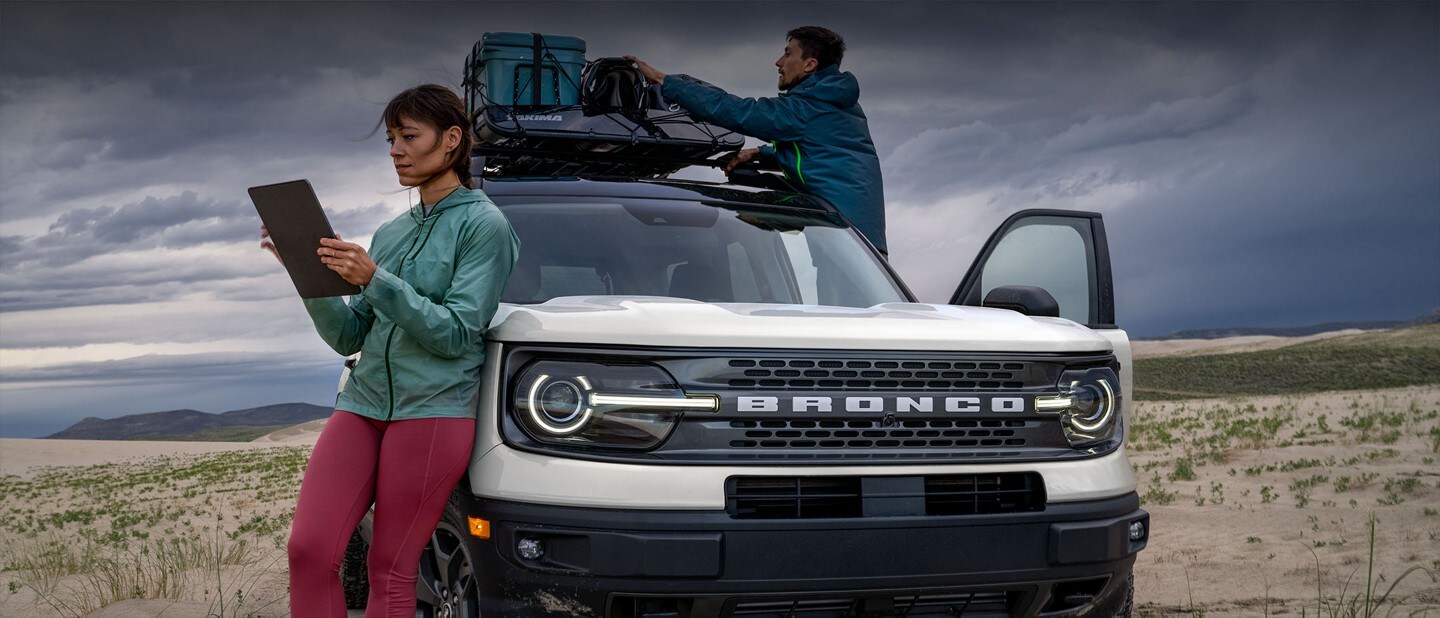 A woman uses a tablet computer while a man packs the roof rack of a white 2023 Ford Bronco® Sport in a desert setting