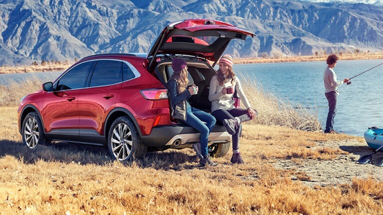 Two women sitting on the trunk of a red 2023 Ford Escape by a lake and a man fishing.