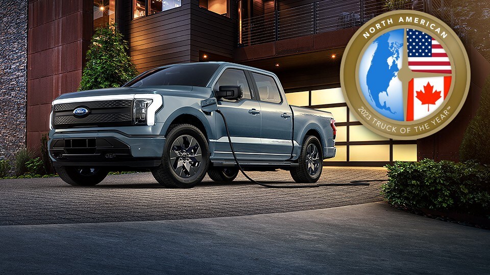 North American 2023 Truck of the Year TM. A silver/blue 2023 Ford F-150 Lightning charging outside of a house.