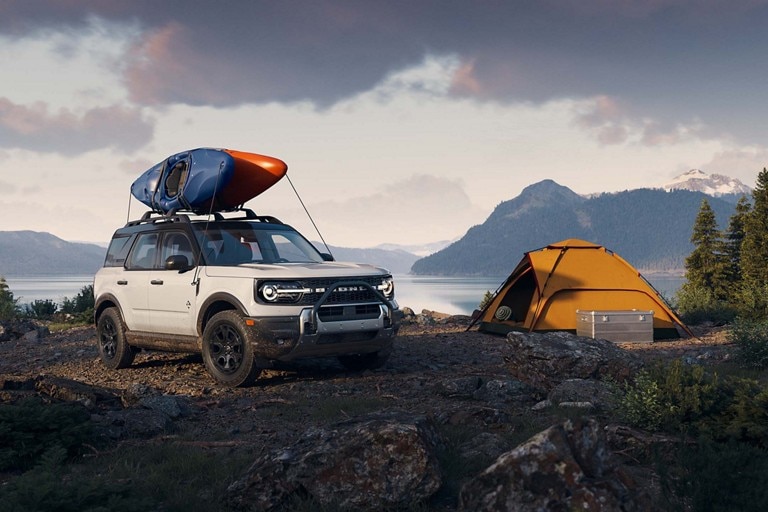 A white Bronco Sport with a canoe on its roof is parked near a yellow tent.