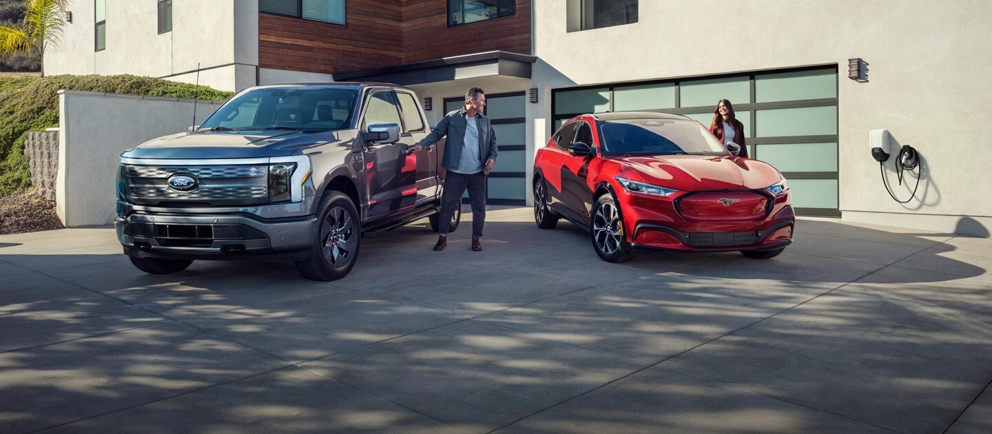 Two people open the driver doors of a gray 2023 Ford F-150 Lightning and red 2023 Ford Mustang Mach-E parked in the driveway.