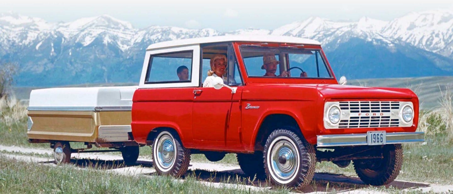 Family in a 1966 Ford Bronco® Sport Wagon in Royal Maroon with Wimbledon White roof towing a camper