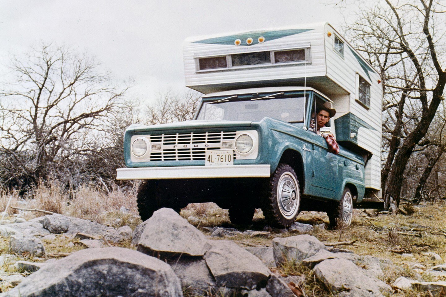 A 1967 Ford Bronco in Peacock Blue with a Camper Body being driven over rocky terrain