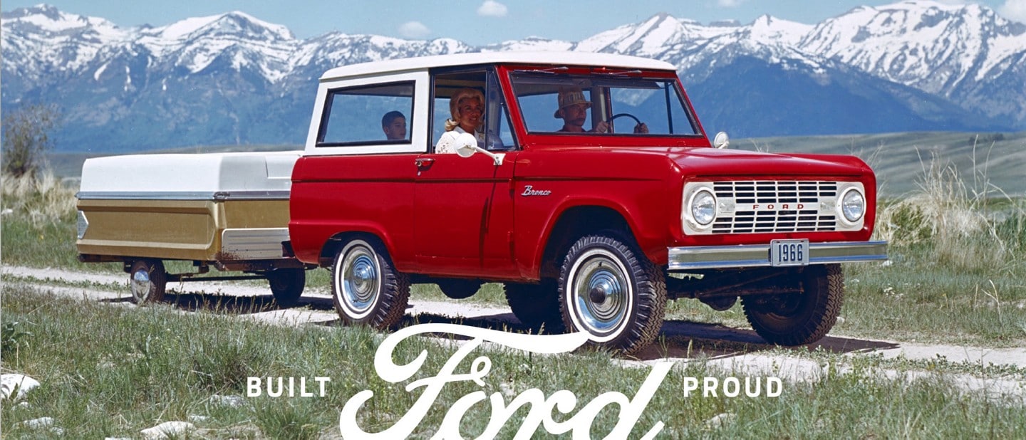 A family riding in a 1966 Ford Bronco Sport Wagon in Royal Maroon with Wimbledon White roof pulling a pop up camper trailer on a two trail in the mountains