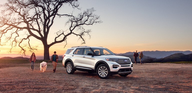 A family with a dog approaches a white Ford Explorer parked under a tree.