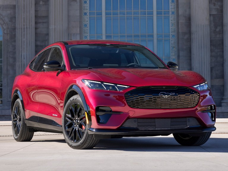A blue Ford Mustang Mach-E® SUV being driven on a tree-lined country road surrounded by greenery.