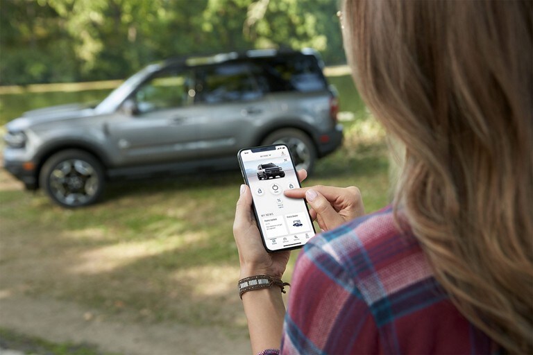 Woman looking at FordPass™ App on mobile phone with 2023 Ford Bronco® Sport SUV in the background