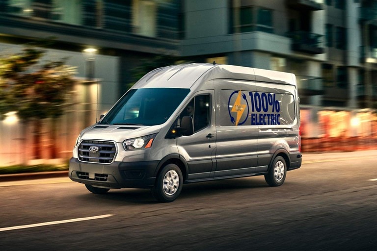 A 2023 Ford E-Transit™ van being driven down a city street at dusk