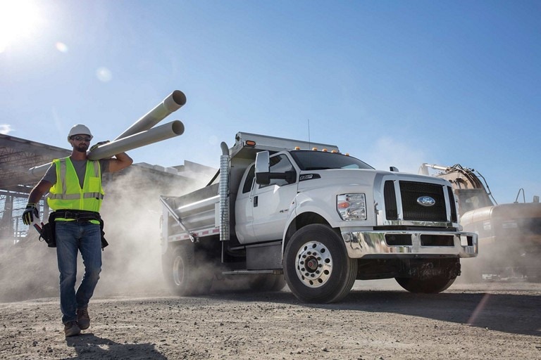 Worker walking on a dusty construction site near a 2026 Ford Medium Duty truck with dump upfit shown in Oxford White