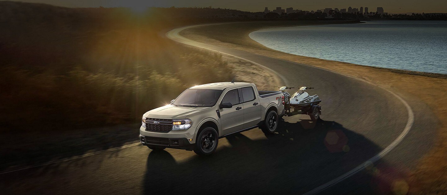 A 2024 Ford Maverick® in Terrain with jet skis being towed on a city highway at sunset near water
