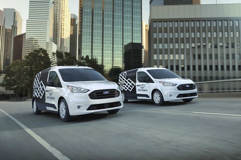 Two 2023 Ford Transit Connect Cargo Vans in Frozen White with aftermarket business graphics being driven on a city street