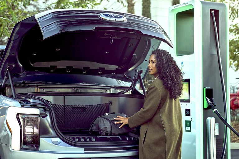 Woman stands in front of an F-150 Lightning with an open frunk at a public charging station.
