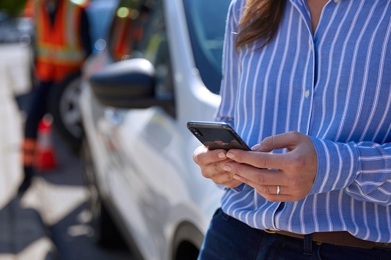 A woman uses a mobile phone for roadside assistance while a tow truck driver oads her car in the background