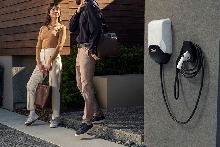 A couple stand near the entrance of a house next to a Ford charger installed on the wall