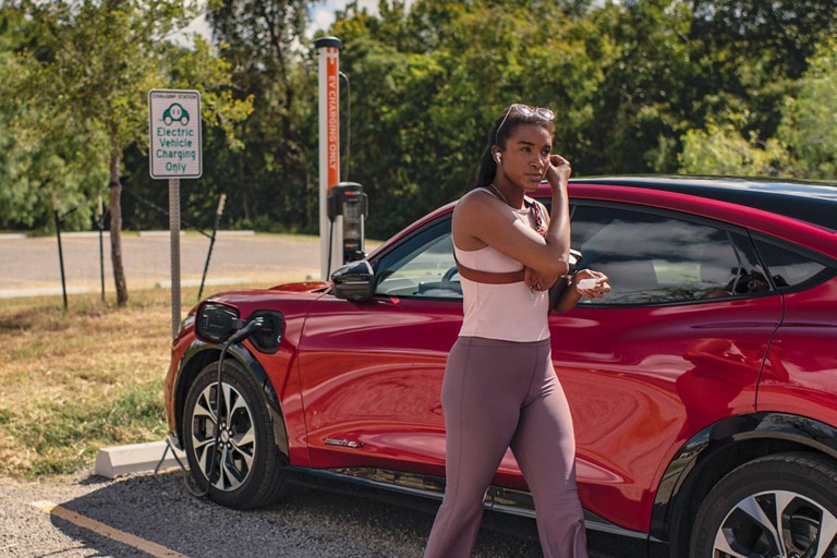 A woman walks away from a red mach E that is plugged into a public charger.