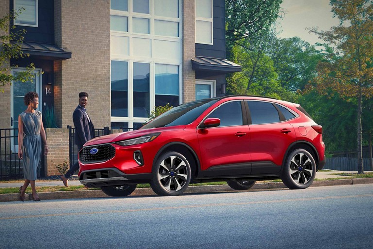 Two people stand near a red Ford Escape parked on a city street.