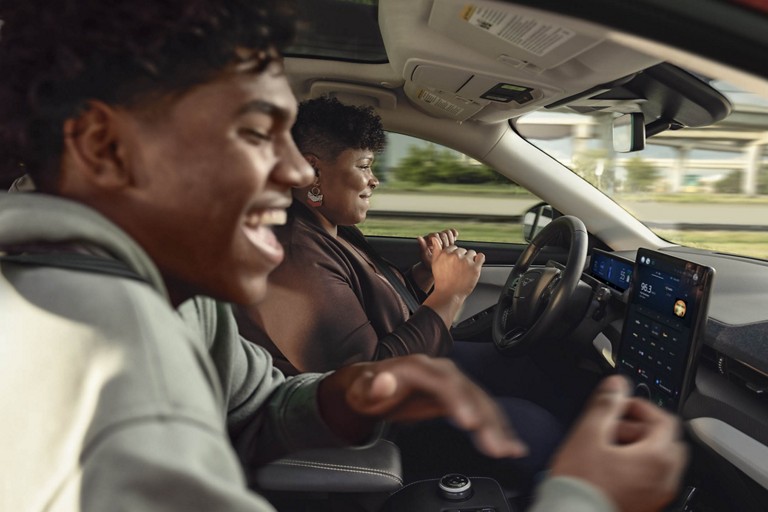 Interior vehicle view of a driver and passenger laughing together while the driver’s hands are off the wheel.