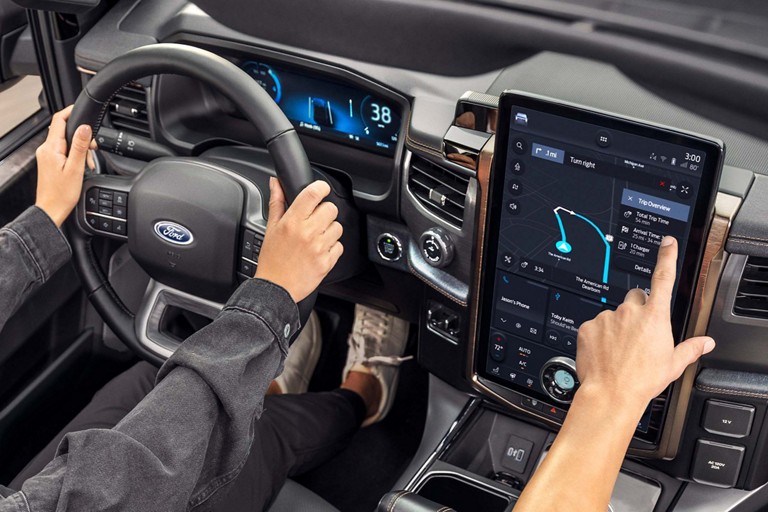 Person driving a Ford vehicle while a passenger interacts with the Connected Navigation screen.