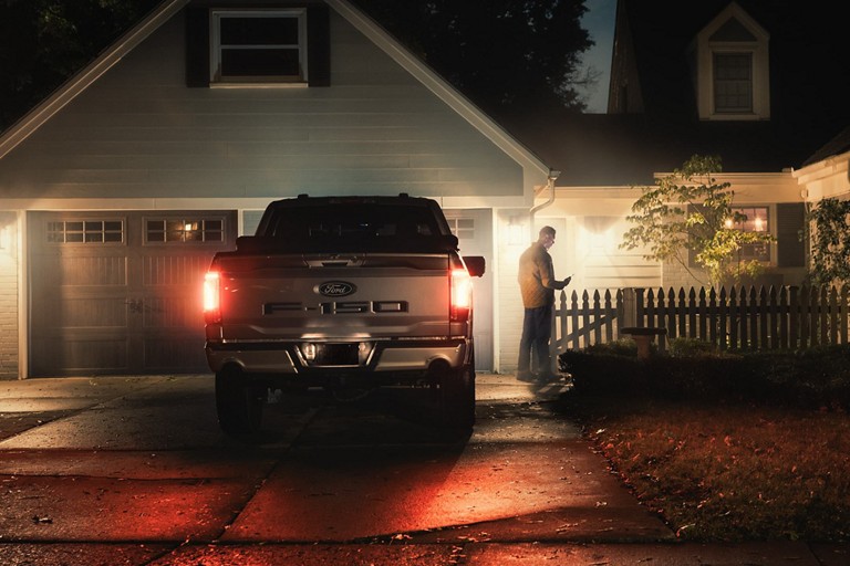 Person holding a smartphone standing next to an F-150 Lightning in a residential driveway. 