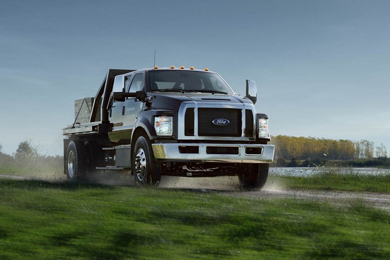 2025 Ford Medium Duty truck shown in Agate Black driving on a gravel road next to a lake