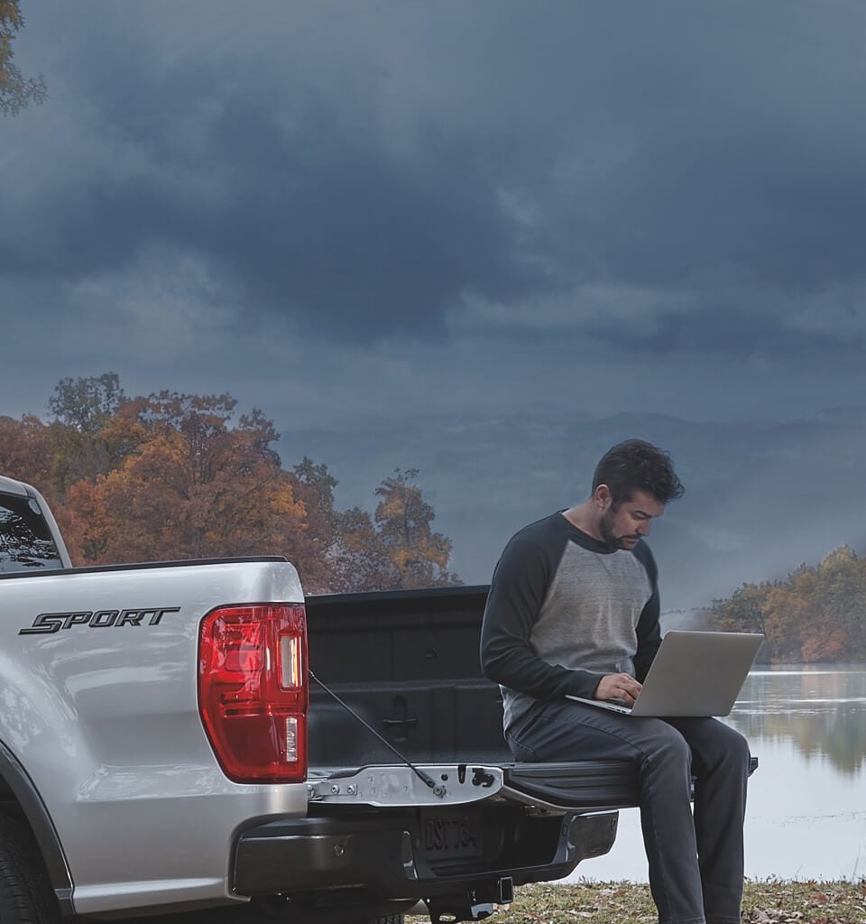 Man using laptop while sitting on truck bed of 2020 Ford Ranger Lariat parked near lake