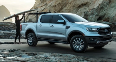 Man placing a surfboard onto the roof rack of a 2020 Ford Ranger LARIAT parked on the sand near a rocky beach