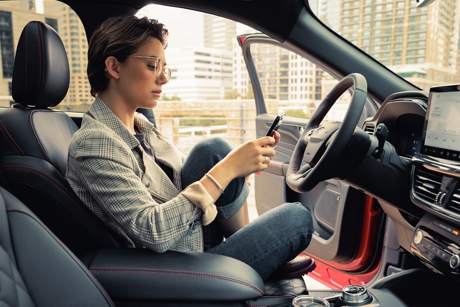 A woman sitting in the driver's seat of a parked red Ford vehicle with the door open. She is using her smartphone