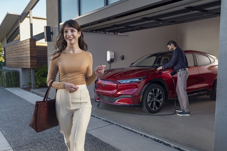 A woman walking away from a red Ford Mustang Mach-E SUV parked in a garage, while a man plugs a charging cable into the vehicle's port