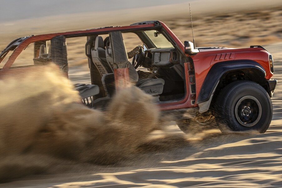 A red 2023 Ford Bronco Raptor with its doors removed is driven across sand dunes.