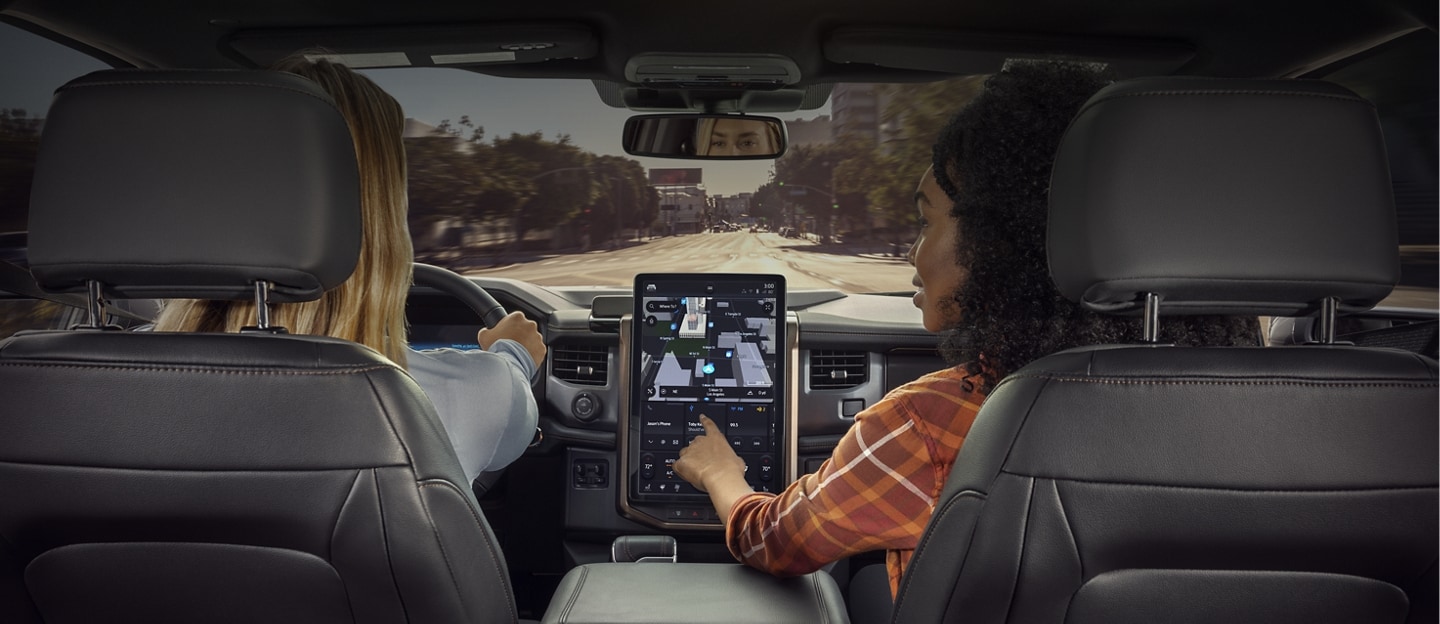 Woman driving a Ford vehicle while a passenger interacts with the Connected Navigation screen.