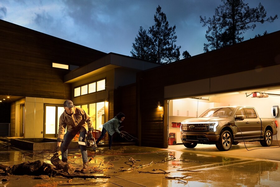 Couple cleaning up storm debris in a driveway with a Ford F-150® Lightning™ in the garage hooked up to charger