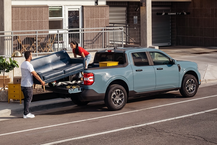 A couple unloads a couch from the bed of a light blue Ford Maverick.