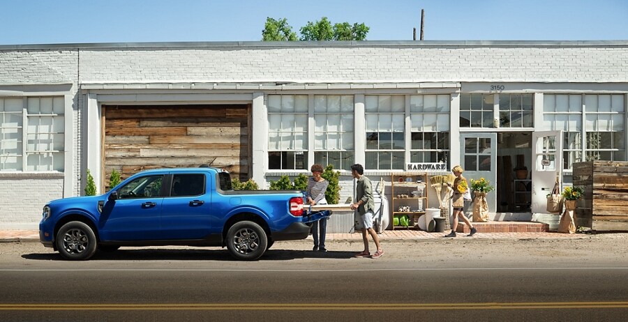 Two people unloading items from the truck bed of a 2026 Ford Maverick® XLT pickup model in Argon Blue parked in front of a building
