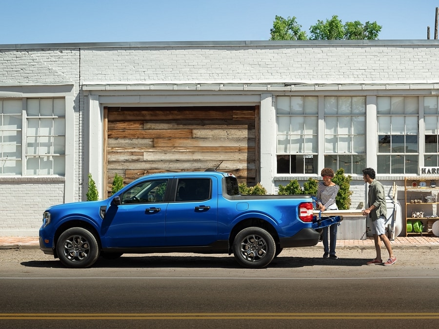 Two people unloading items from the truck bed of a 2026 Ford Maverick® XLT pickup model in Argon Blue parked in front of a building