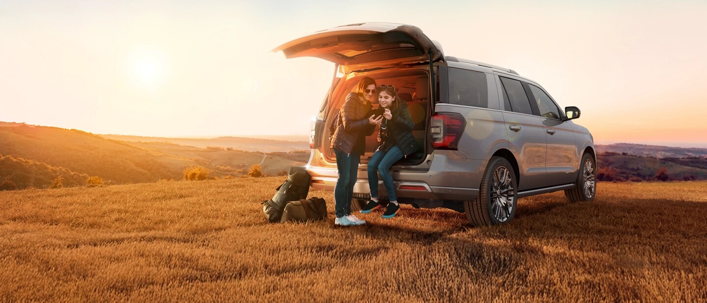 Two people sit in the tailgate of a Ford Expedition parked in an empty field and look together at a phone screen.