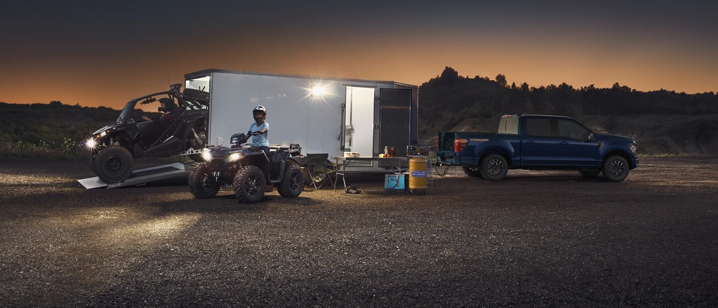 Two people drive ATVs from a trailer hitched to a parked F-150.