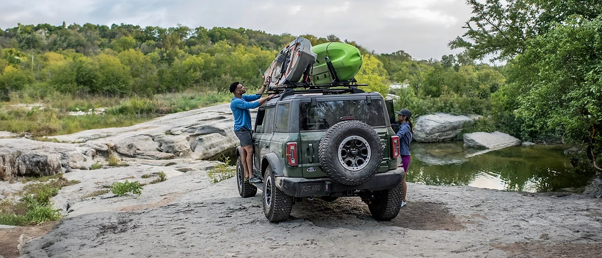 A man unloading an inflatable raft from the roof of a 2025 Ford Bronco® SUV parked on rocks near a stream