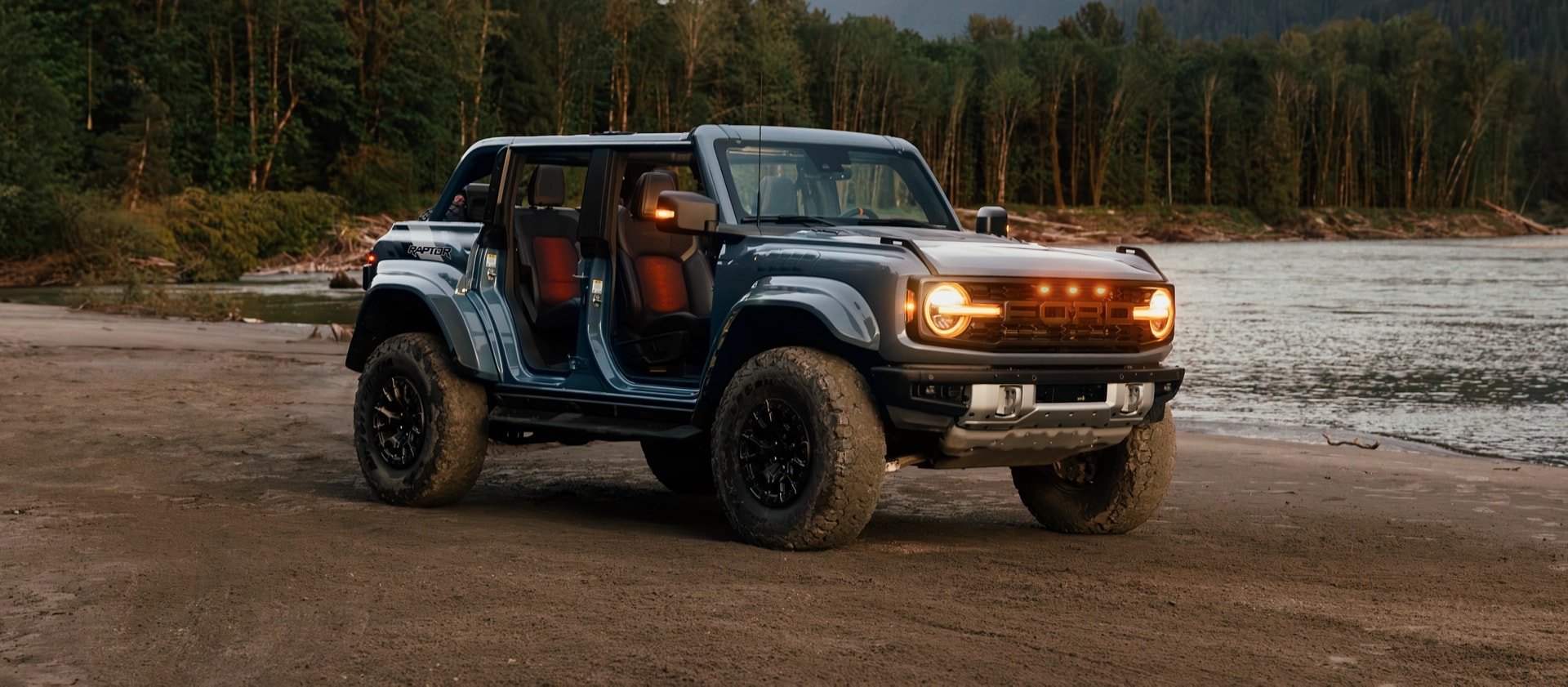 A 2025 Ford Bronco® SUV with the doors off and lights on, parked near a lake in the forest