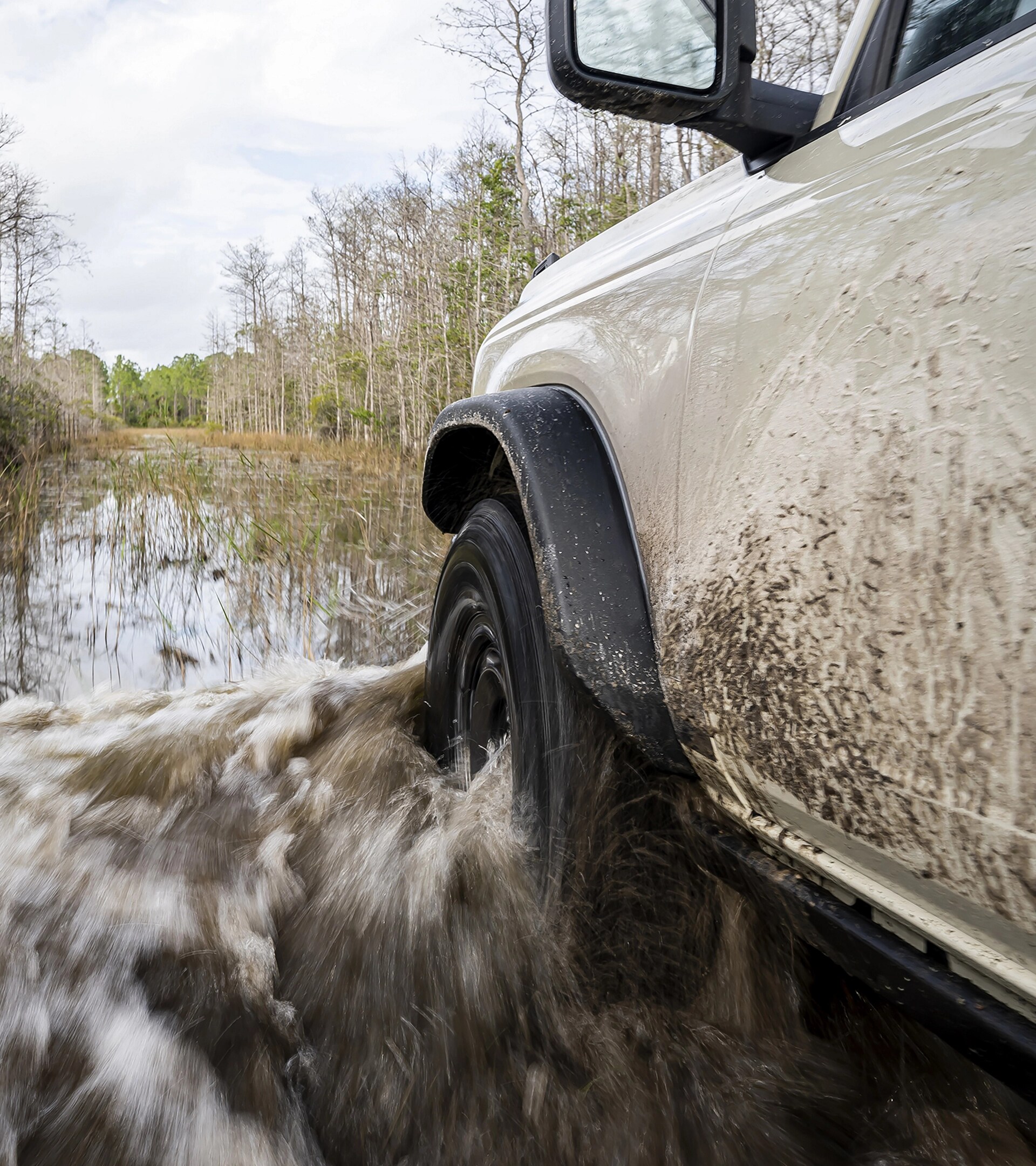 A close-up of the front driver's-side wheel and fender of a 2026 Ford Bronco® SUV as it's driven through a river