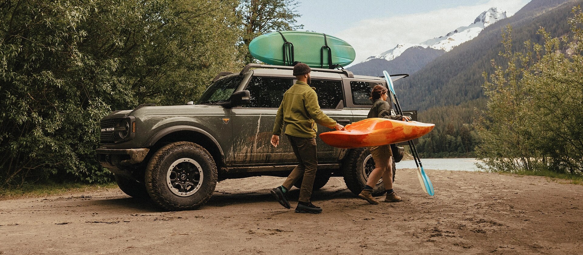 A man and woman carrying a kayak and paddle away from a 2026 Ford Bronco® SUV parked on a mud flat