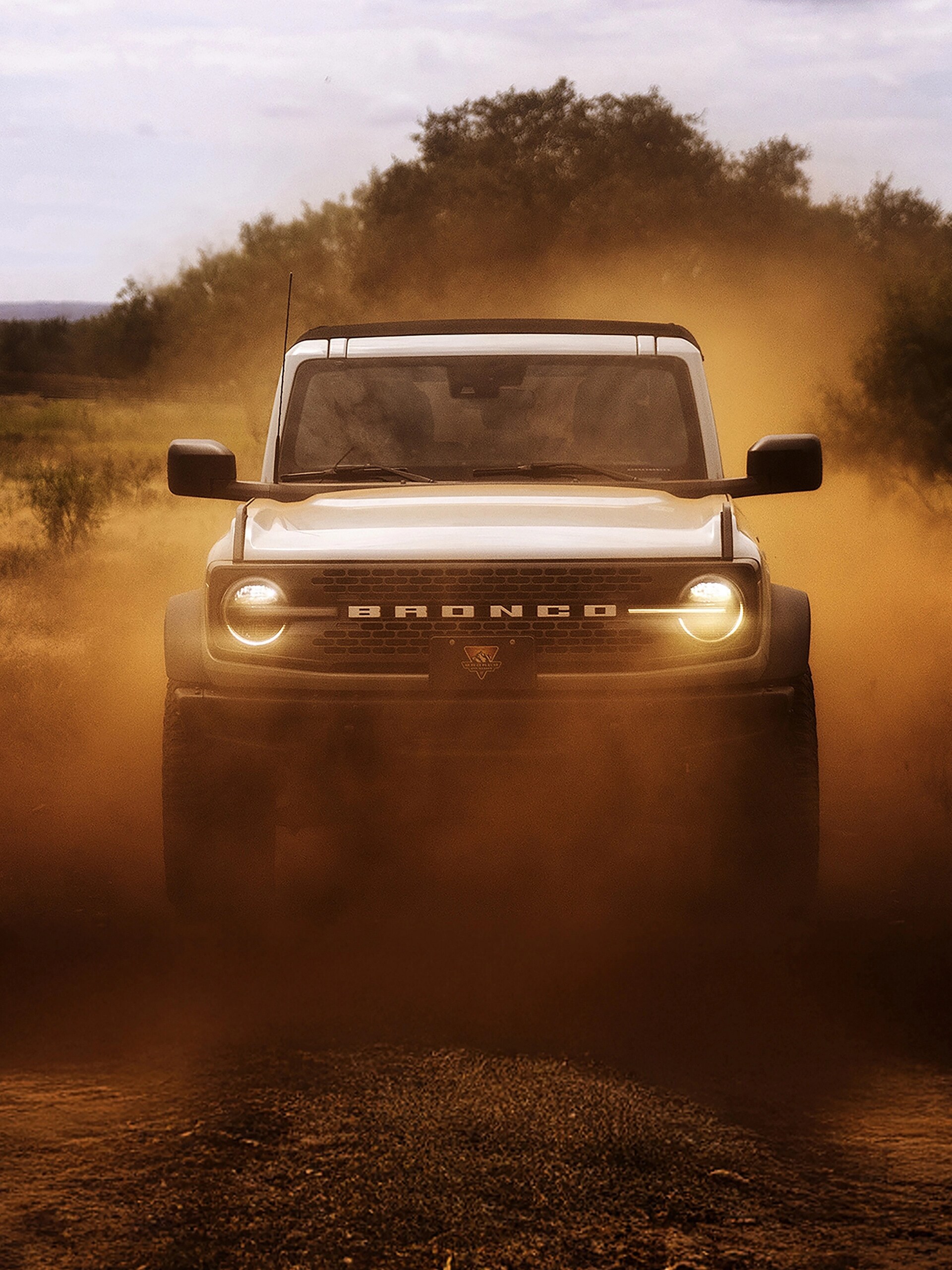 The front of a 2026 Ford Bronco® SUV, its headlights shining through a cloud of red dust